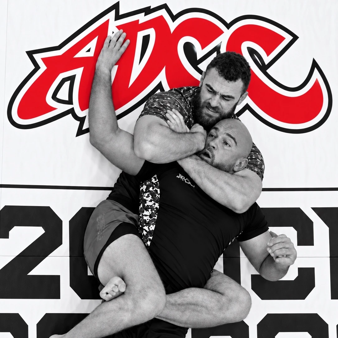 Two grapplers locked in a submission exchange in front of the ADCC logo