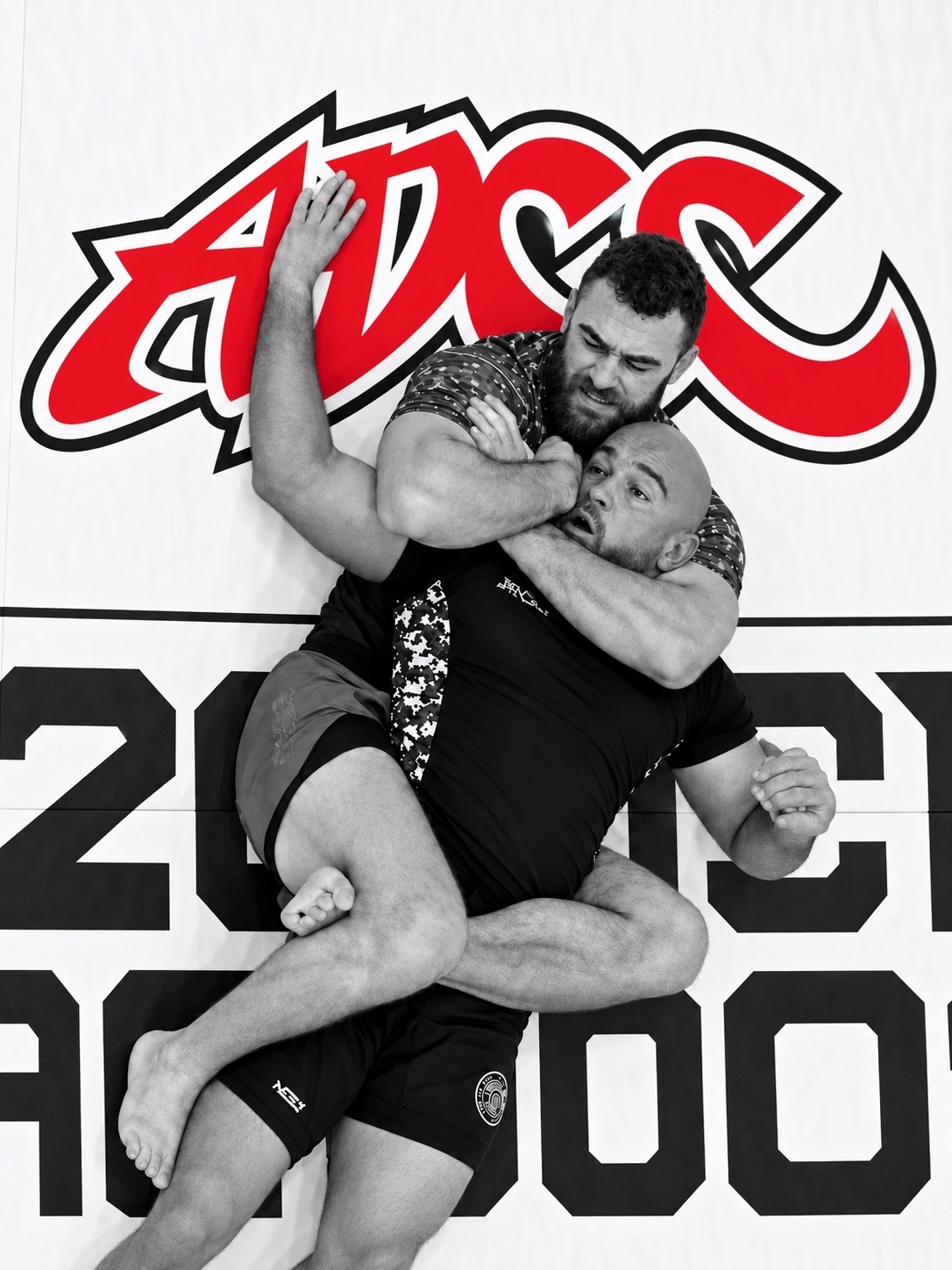 Two grapplers locked in a submission exchange in front of the ADCC logo